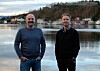 To menn står ved ein sjø med busetnad og skogkledde åsar i bakgrunnen.