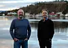 To menn står ved ein sjø med busetnad og skogkledde åsar i bakgrunnen.