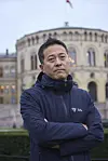 Mann i mørk LHL-jakke står med armane i kors framfor ein utsmykka bygning.