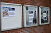 Tre innramma oppslagstavler med plakatar og informasjon på ein raud teglsteinsvegg.