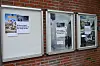 Tre innramma oppslagstavler med plakatar og informasjon på ein raud teglsteinsvegg.