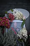 Blomsterbukettar og grøne plantar framfor eit innramma portrett av kong Harald.