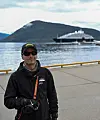 Ein person står på kaia framfor ei stor luksusyacht med fjell i bakgrunnen.
