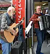 Ein gitarist og ein trekkspelar spelar utanfor ein butikk medan folk ser på.
